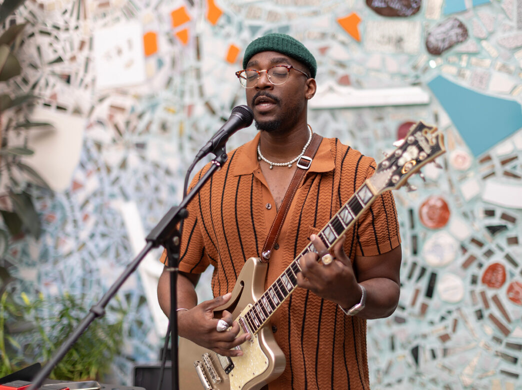 A musician wearing a green knit cap and glasses plays the electric guitar and sings into a microphone in of a mosaic-filled courtyard.