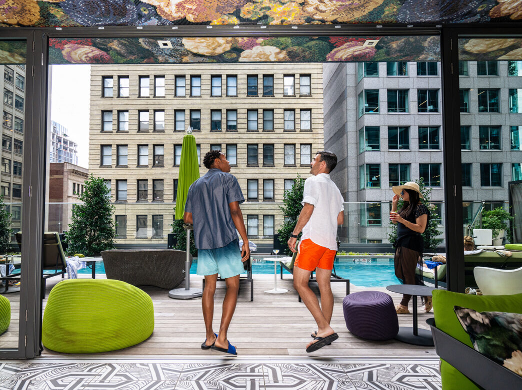 Los huéspedes, vestidos con trajes de baño, pasean y se relajan en la terraza de la alberca de la azotea del W Philadelphia, rodeados de sillones, una decoración colorida y vistas a los rascacielos de Center City.