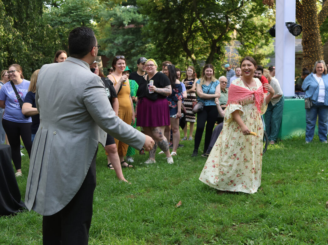 Guests dressed in period-inspired attire laugh and interact during a Zoo Done It mystery event at the Philadelphia Zoo, as a crowd of attendees watch the performance on a grassy lawn.