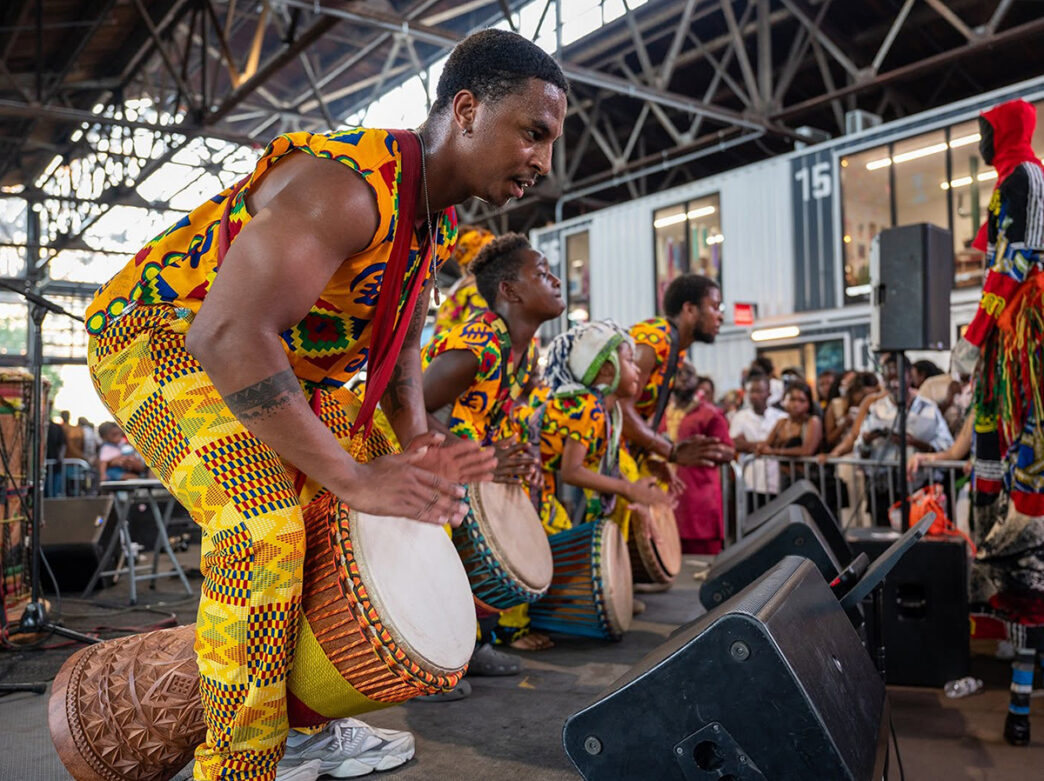 Performers in brightly patterned West African attire play hand drums on stage during the ACANA Festival at Cherry Street Pier, entertaining a crowd gathered inside the venue.