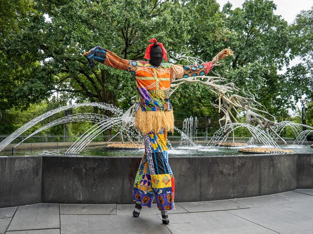 A performer in colorful patchwork costume dances in front of a spraying fountain decorated at the African Culture Fest at the Philadelphia Zoo.