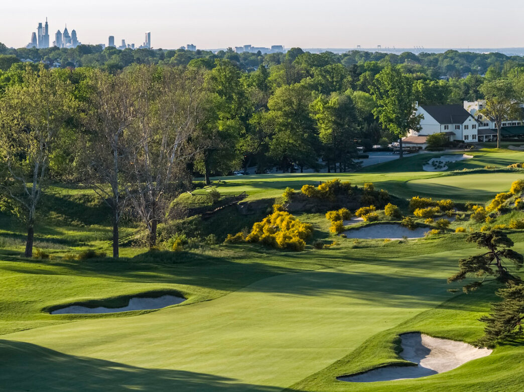Rolling green fairways and sand bunkers at Merion Golf Club with green trees and the Philadelphia skyline in the distrance.
