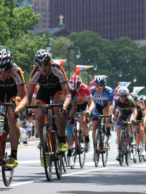 A pack of professional cyclists wearing helmets, sunglasses and cycling jerseys races down the Benjamin Franklin Parkway during the Philadelphia Cycling Classic.