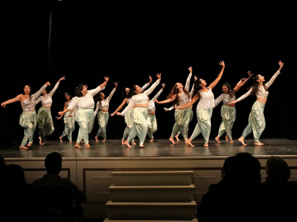 A group of dancers in shimmering silver tops and flowing green pants perform a choregraphed dance routine on stage in front of a live audience.