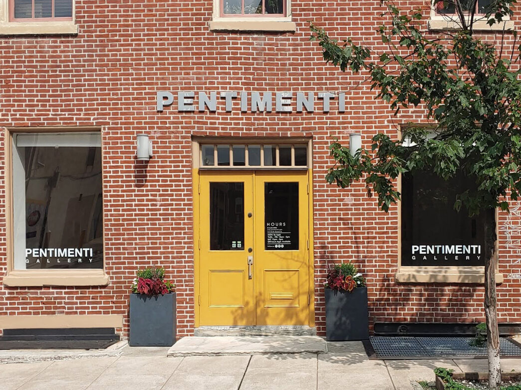 A set of bright yellow doors are centered between two large windows on the red brick building of PENTIMENTI.