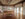 Sunlight fills a spacious gallery at Al-Bustan Seeds of Culture, featuring hardwood floors, exposed wood beams, large industrial style windows, framed artwork displayed on white walls and a cushioned bench in the center of the space.