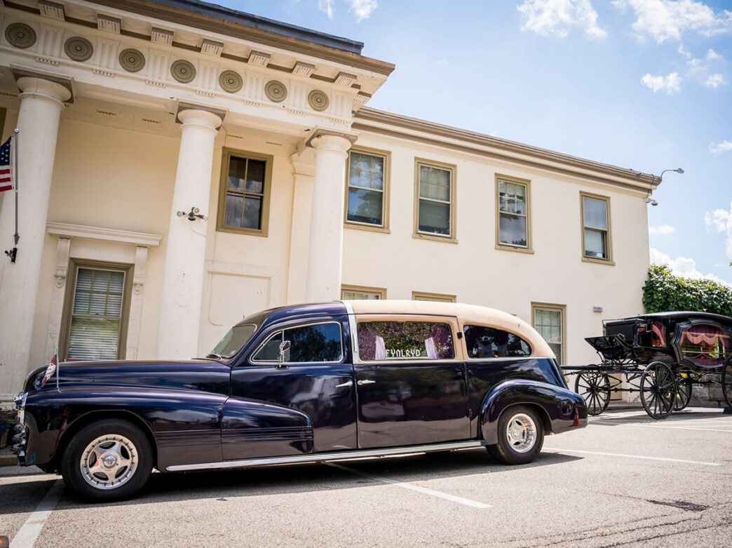 A vintage black-and-cream hearse is parked outside a white gatehouse during the annual Hearse Show, with a horse-drawn carriage displayed behind it.
