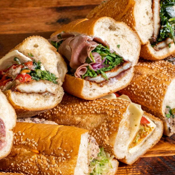 Una variedad de sándwiches recién hechos apilados sobre una mesa de madera, en la que se pueden apreciar los sándwiches rellenos de cerdo asado, filetes de pollo, carne de res asada, embutidos, quesos y verduras en panecillos con semillas.