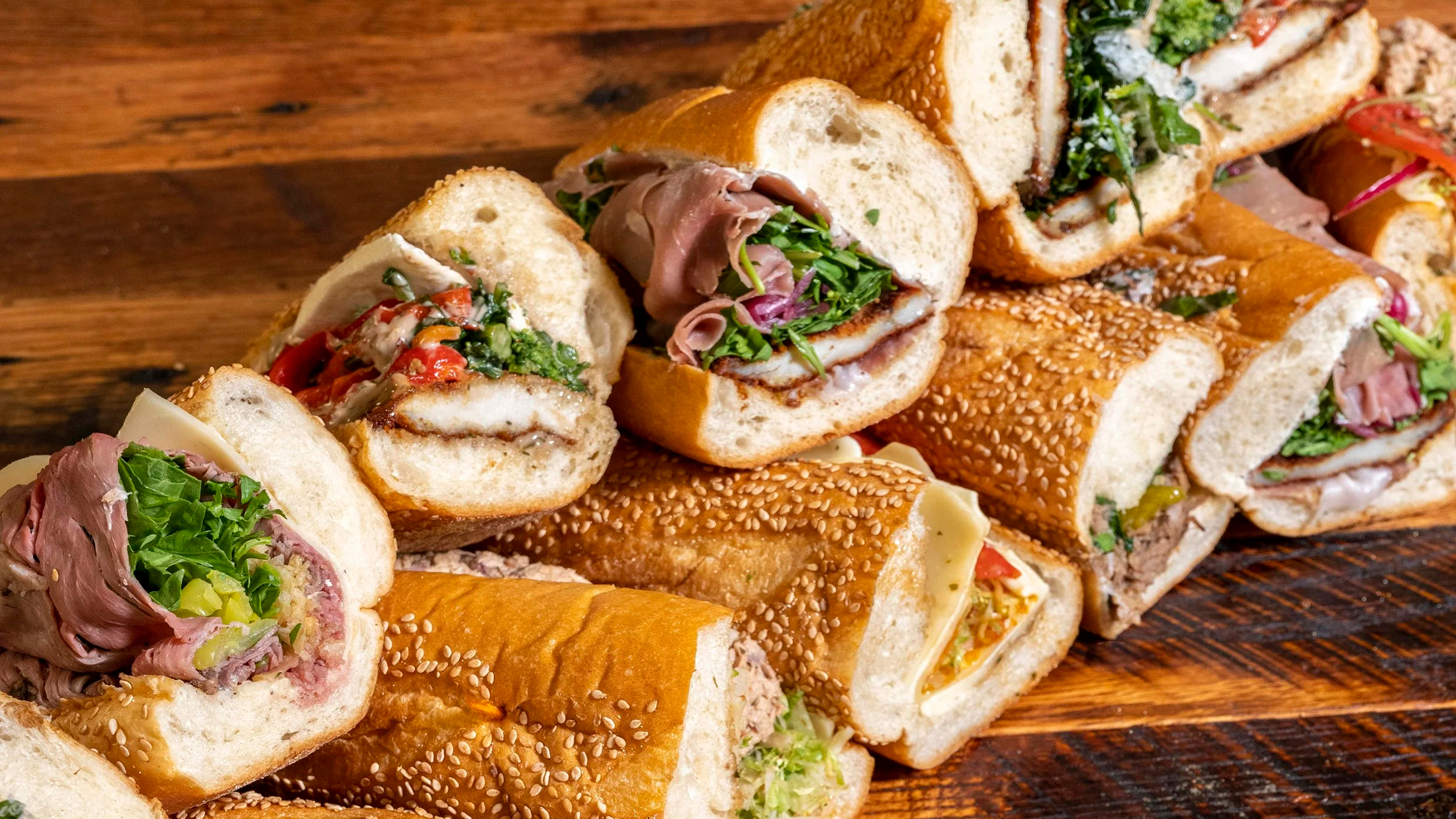 An assortment of freshly made hoagies stacked on a wooden table, showcasing the hoagies filled with roasted pork, chicken cutlets, roast beef, meats, cheeses, vegetables on seeded rolls.