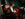 A jazz trio performs inside a bar, with a trumpet player, upright bassist and drummer playing in front of a mirror on a dark green wall.
