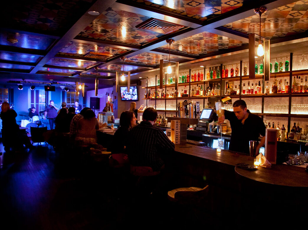 A bartender pours a cocktail behind the bar as guests enjoy drinks and a live band performs on a small stage at The Twisted Tail in Philadelphia.