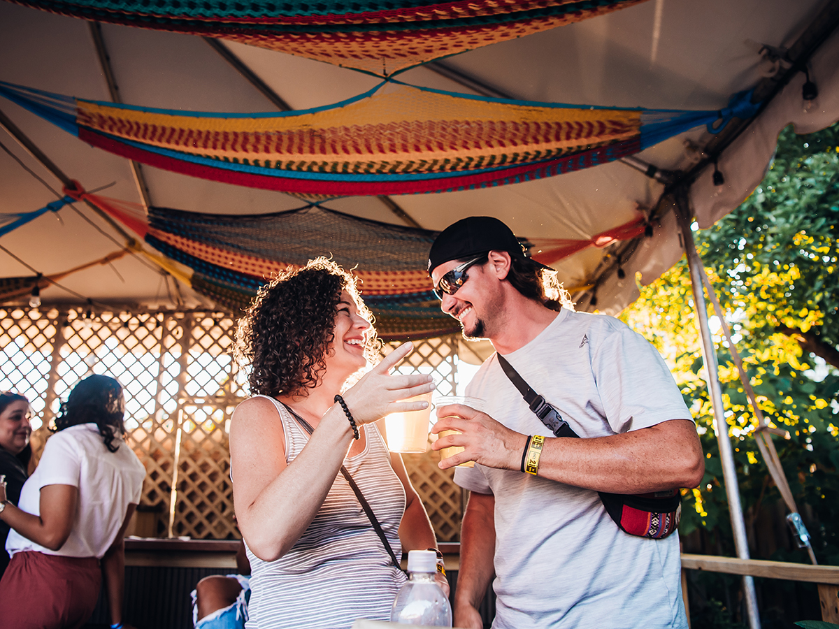 Two visitors laugh and enjoy drinks beneath a colorful woven hammocks hanging from the ceiling of a party tent during golden hour.