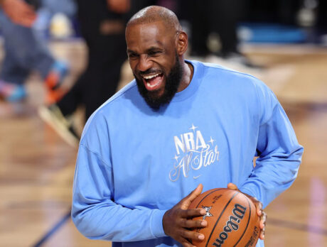 LeBron James smiles while holding a basketball and wearing a light blue NBA All Star crewneck.