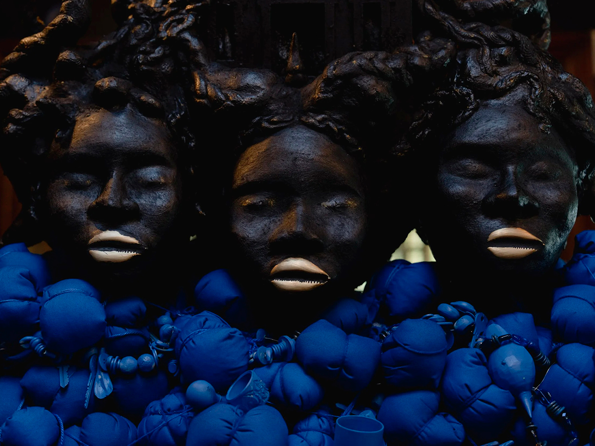 A close-up of Oracle by artist Vanessa German, featuring three sculpted faces with gold lips surrounded by vivid blue fabric, beads and found objects.