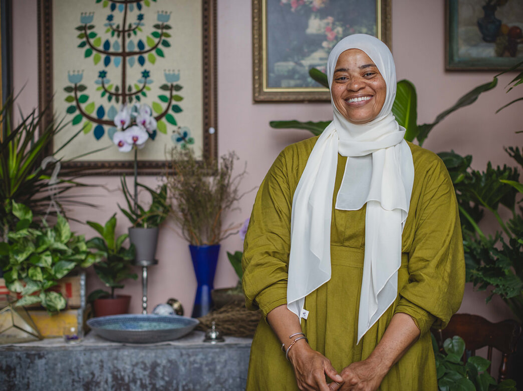 A smiling woman wearing a white hijab and an olive green dress poses for a photo while surrounded by lush houseplants, floral artwork and botanical decor.