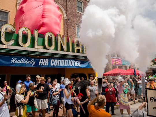 Crowds gather outside The Colonial Theatre during Phoenixville's BlobFest as participants in colorful costumes spray fire extinguishers into the air during the festival's signature parade, with the theater's iconic marquee in the background.