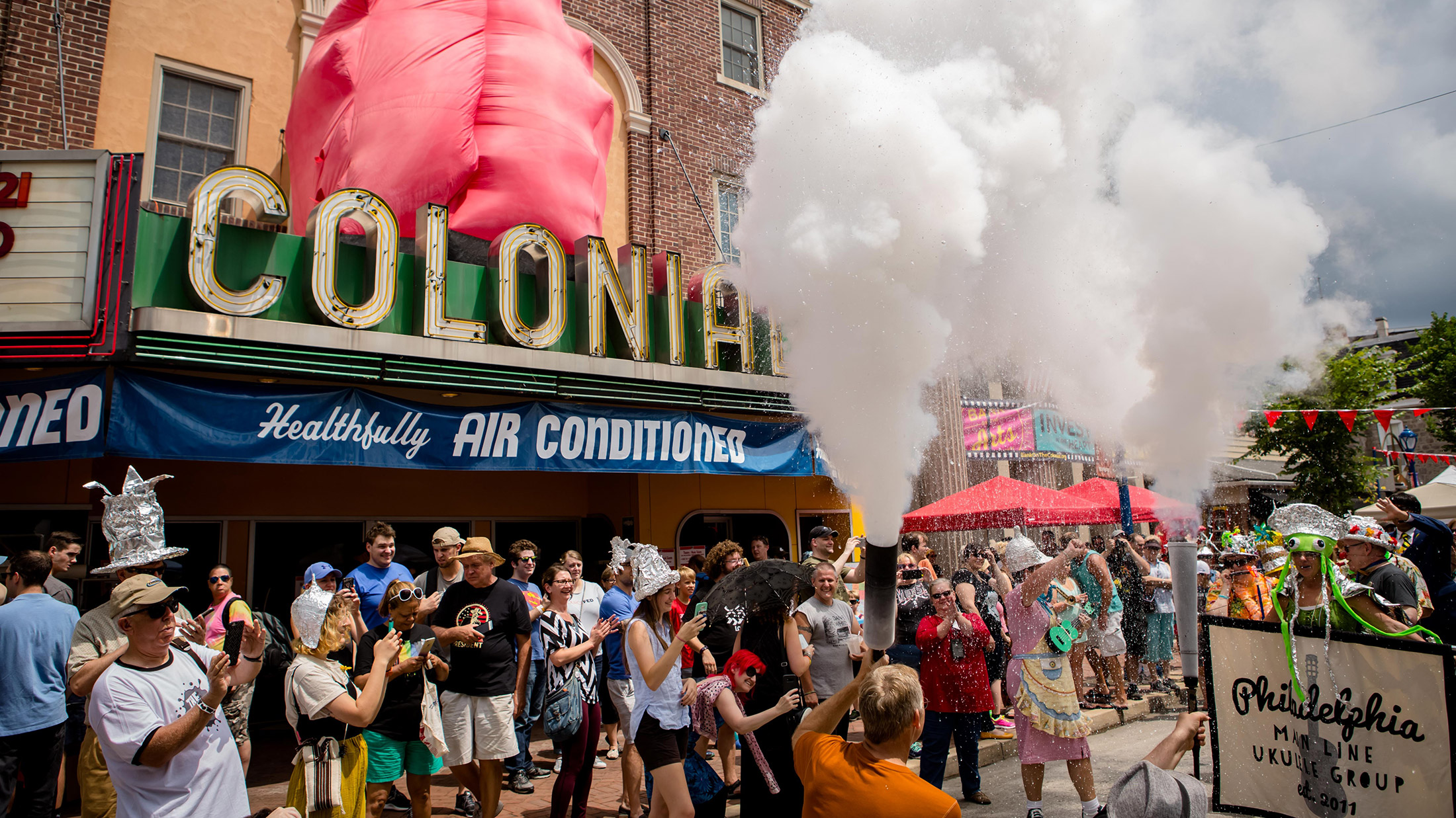 Crowds gather outside The Colonial Theatre during Phoenixville's BlobFest as participants in colorful costumes spray fire extinguishers into the air during the festival's signature parade, with the theater's iconic marquee in the background.