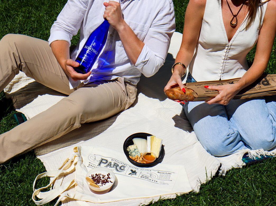 Par un après-midi ensoleillé, un couple est assis sur une couverture de pique-nique blanche dans l'herbe, avec du pain, du fromage, de l'eau pétillante et des en-cas achetés chez Parc.