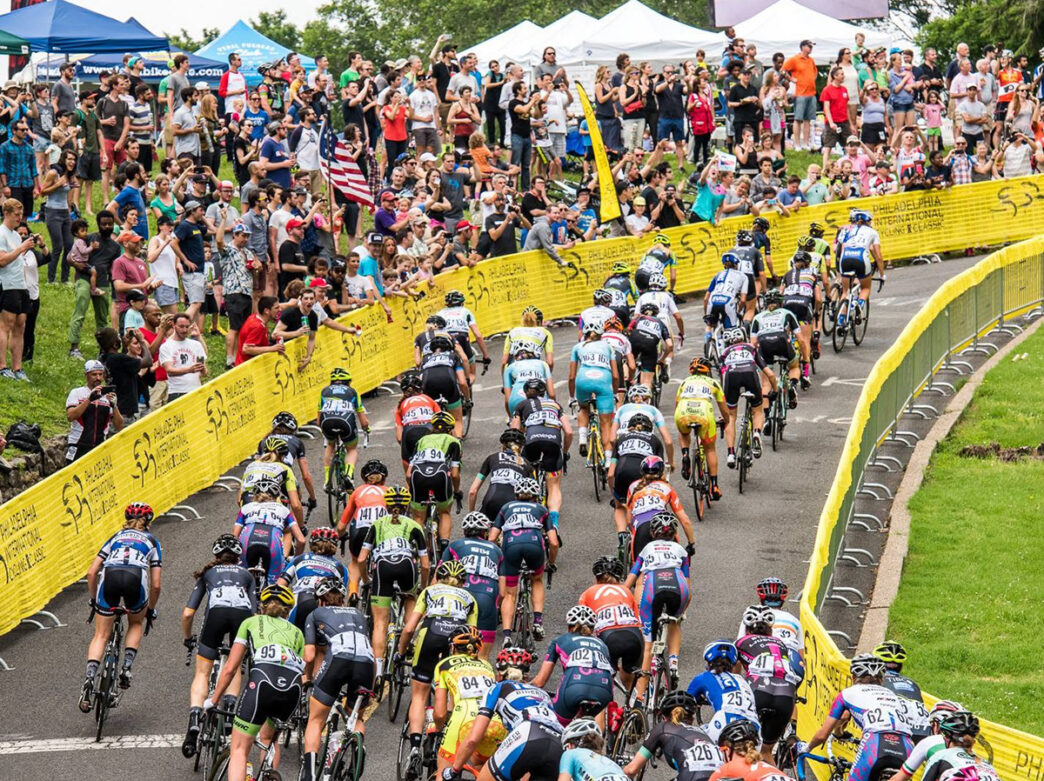 Spectators standing behind a yellow fence cheer and watch as professional cyclists race by.