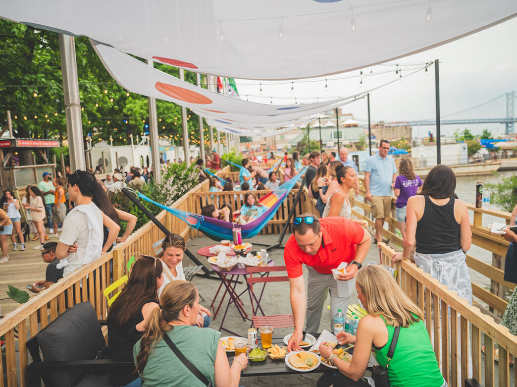 As pessoas se alimentam em uma mesa repleta de comida e bebidas, mulheres estão sentadas em uma rede colorida e uma multidão caminha à beira-mar no Spruce Street Harbor Park.