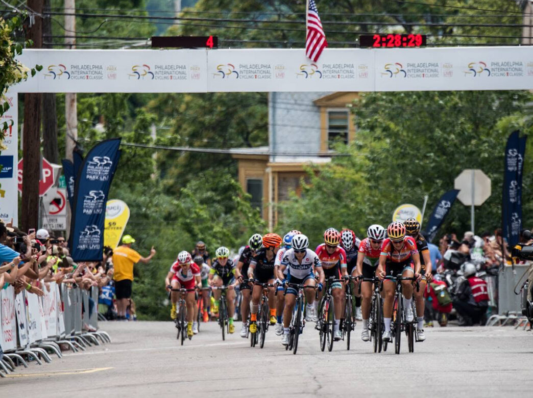 Elite female cyclists race toward the finish line beneath a Philadelphia Cycling Classic banner as crowds line the finish line and cheer them on.