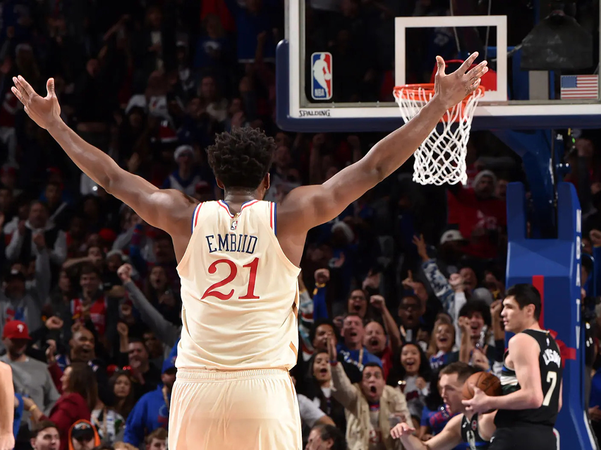Joel Embiid se tient dos à la caméra sous le panier, les bras levés, tandis qu'une foule en délire l'acclame.