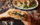 A diner holds a sandwich filled with roasted vegetables and greens over a restaurant table, with a salad and wine glass in the background.