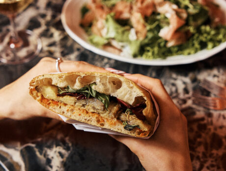 Un comensal sostiene un sándwich relleno de verduras asadas y hojas verdes sobre la mesa de un restaurante, con una ensalada y una copa de vino al fondo.