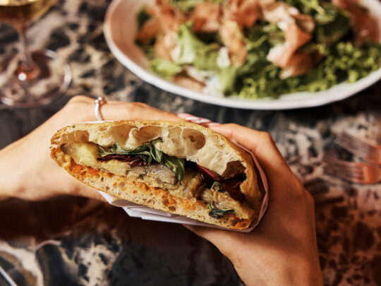 Un comensal sostiene un sándwich relleno de verduras asadas y hojas verdes sobre la mesa de un restaurante, con una ensalada y una copa de vino al fondo.
