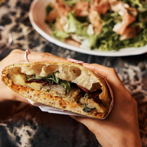 A diner holds a sandwich filled with roasted vegetables and greens over a restaurant table, with a salad and wine glass in the background.