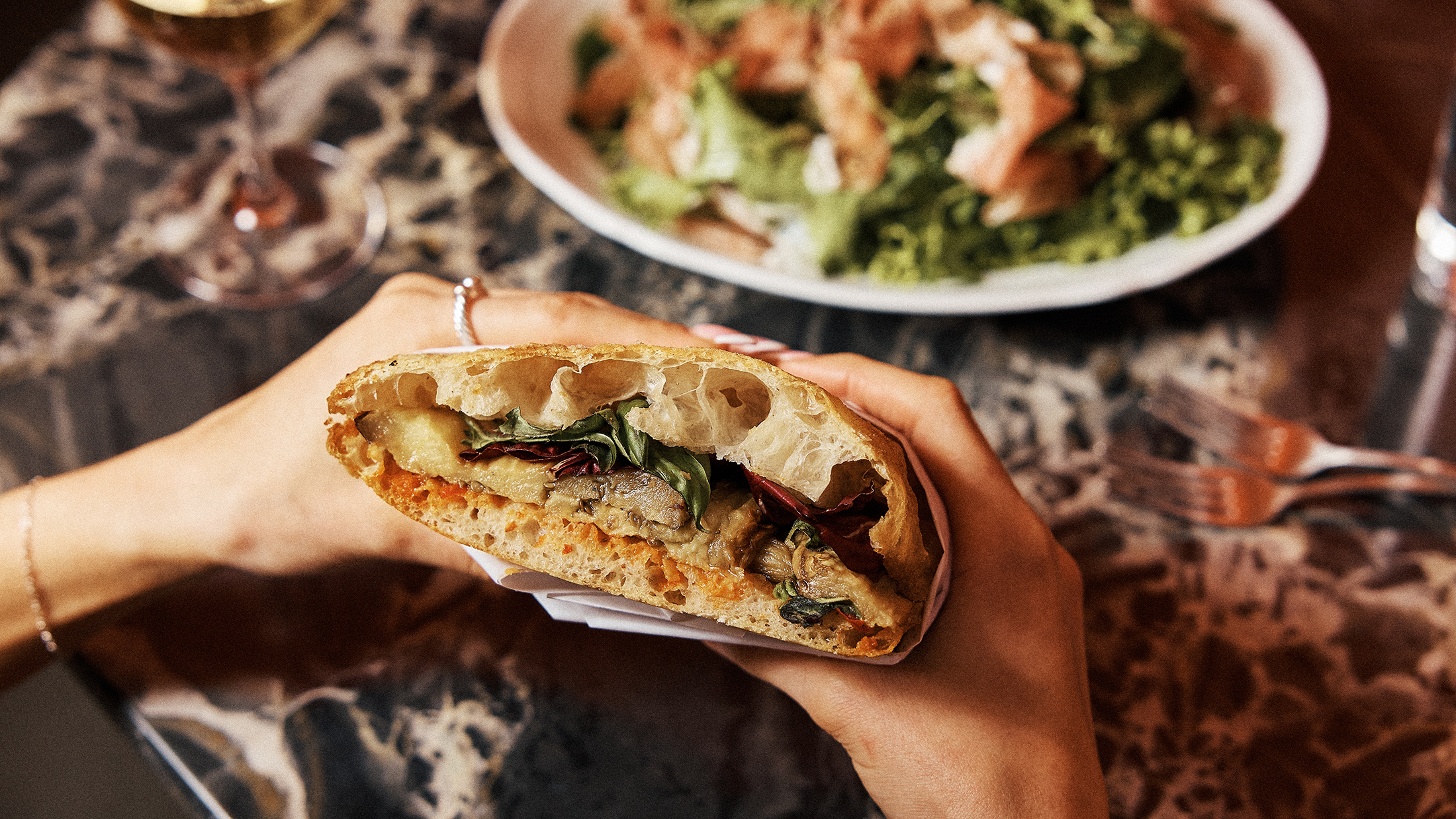A diner holds a sandwich filled with roasted vegetables and greens over a restaurant table, with a salad and wine glass in the background.
