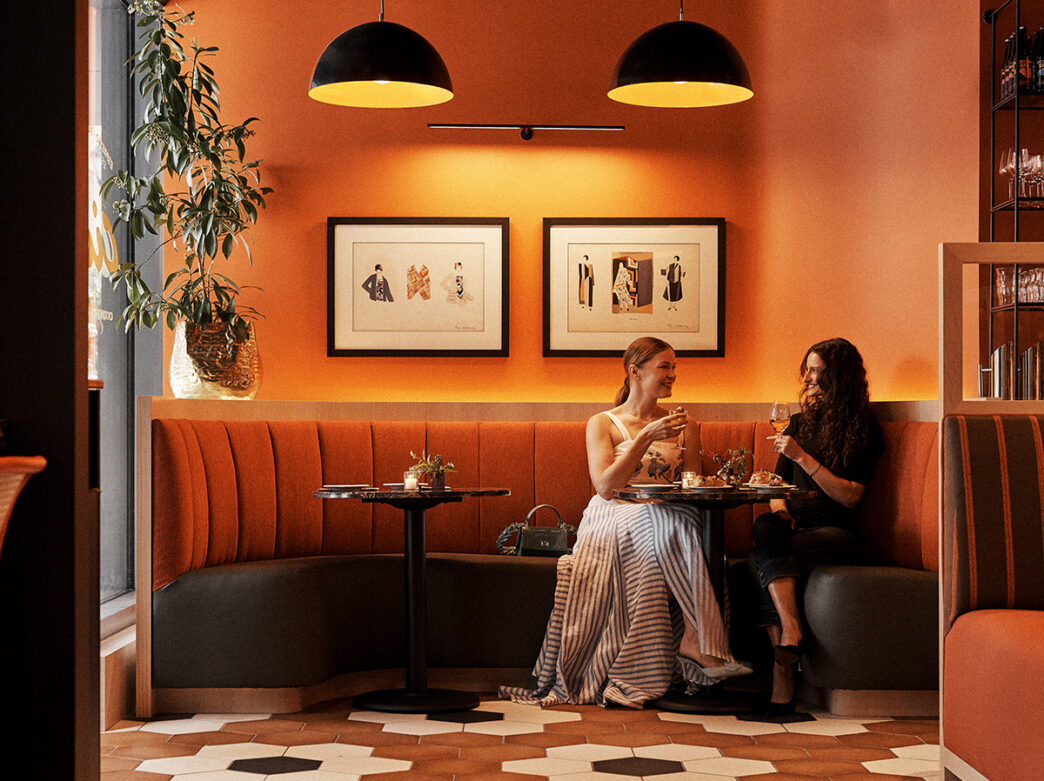 Two women smile and raise glasses together while seated in a cozy orange booth at Bar Cicci, surrounded by warm lighting and framed artwork.