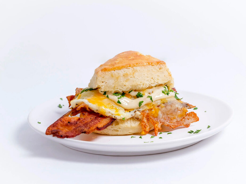 A buttermilk biscuit breakfast sandwich with bacon, eggs and melted cheese sits on a white plate, garnished with chopped parsley.