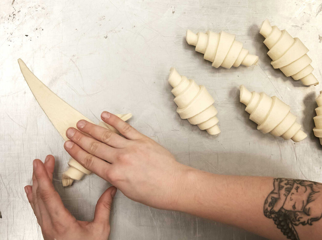 A close-up shows a pair of hands rolling triangular pieces of dough into croissants on a metal work surface.