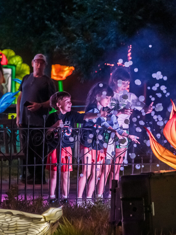Three children play with floating bubbles beside a glowing lotus flower lantern and a swan lantern during the Philadelphia Chinese Lantern Festival at night.