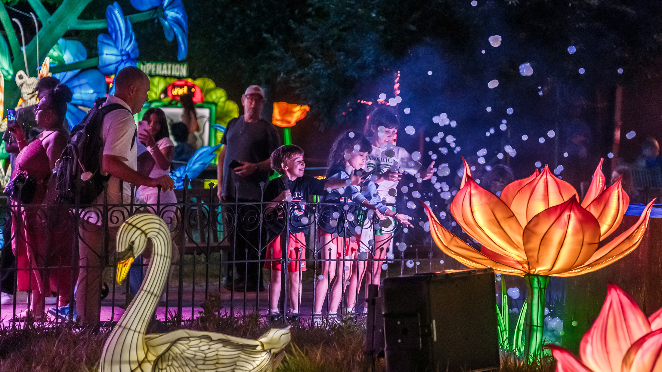 Three children play with floating bubbles beside a glowing lotus flower lantern and a swan lantern during the Philadelphia Chinese Lantern Festival at night.