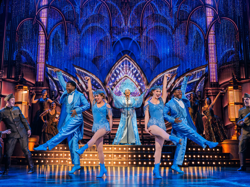 Performers in sparkling blue costumes dance across an elaborate Art Deco-style stage, with a singer at center and ensemble members surrounded them beneath shimmering blue lighting.