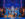 Performers in sparkling blue costumes dance across an elaborate Art Deco-style stage, with a singer at center and ensemble members surrounded them beneath shimmering blue lighting.