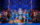 Los artistas, con trajes azules brillantes, bailan en un elaborado escenario de estilo Art Déco, con un cantante en el centro y los miembros del grupo rodeándolo bajo una iluminación azul resplandeciente.