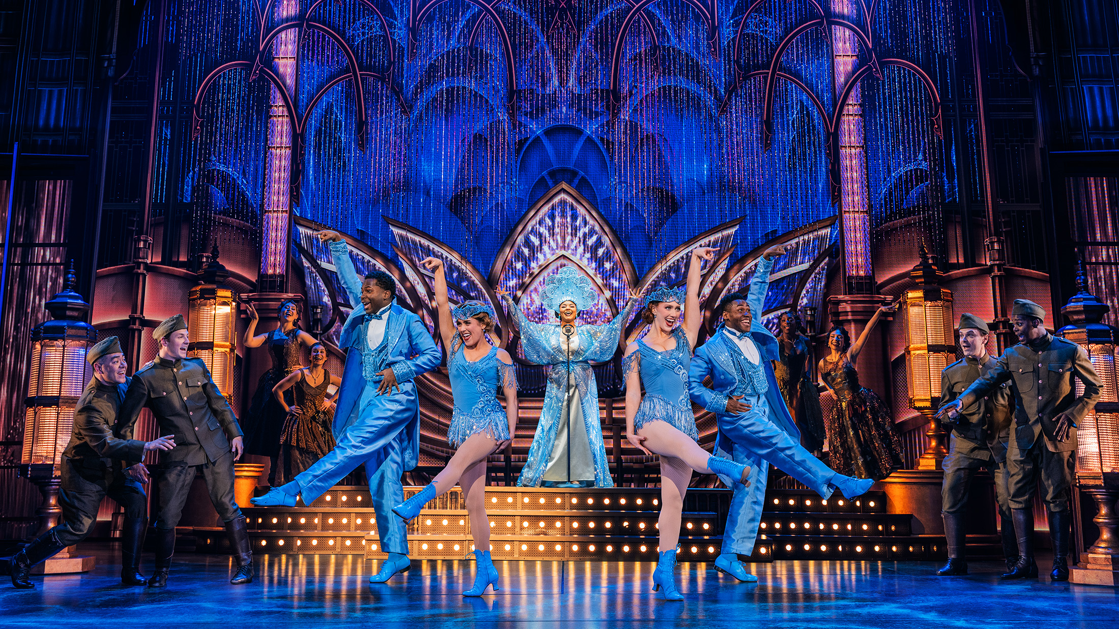 Los artistas, con trajes azules brillantes, bailan en un elaborado escenario de estilo Art Déco, con un cantante en el centro y los miembros del grupo rodeándolo bajo una iluminación azul resplandeciente.