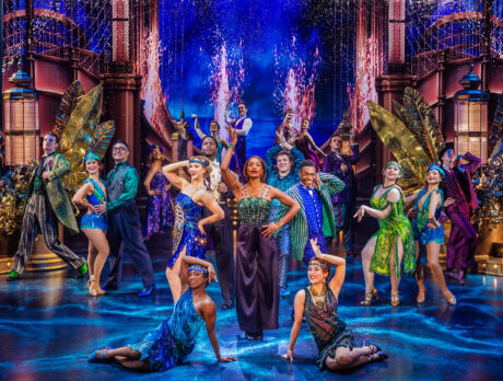 The cast of The Great Gatsby dance and pose on an elaborate Art Deco-inspired stage in colorful, sparkling, 1920s-inspired costumes surrounded by golden set pieces, glowing lights and dramatic cityscape background.