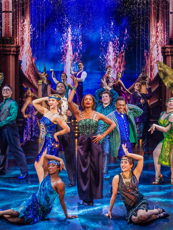 The cast of The Great Gatsby dance and pose on an elaborate Art Deco-inspired stage in colorful, sparkling, 1920s-inspired costumes surrounded by golden set pieces, glowing lights and dramatic cityscape background.
