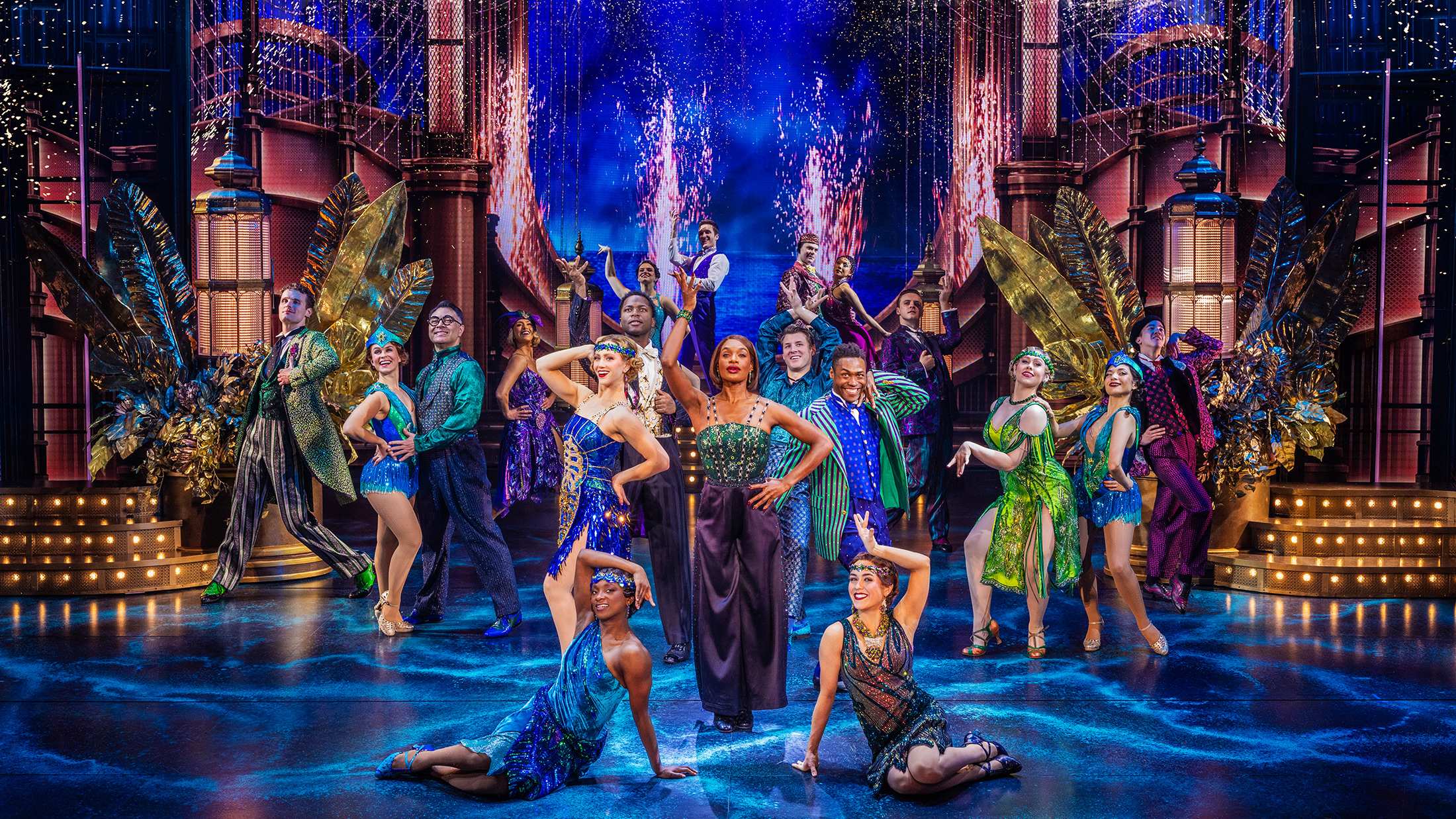 El elenco de *El gran Gatsby* baila y posa en un elaborado escenario de inspiración Art Déco, con trajes coloridos y brillantes inspirados en la década de 1920, rodeados de elementos escenográficos dorados, luces resplandecientes y un espectacular paisaje urbano de fondo.