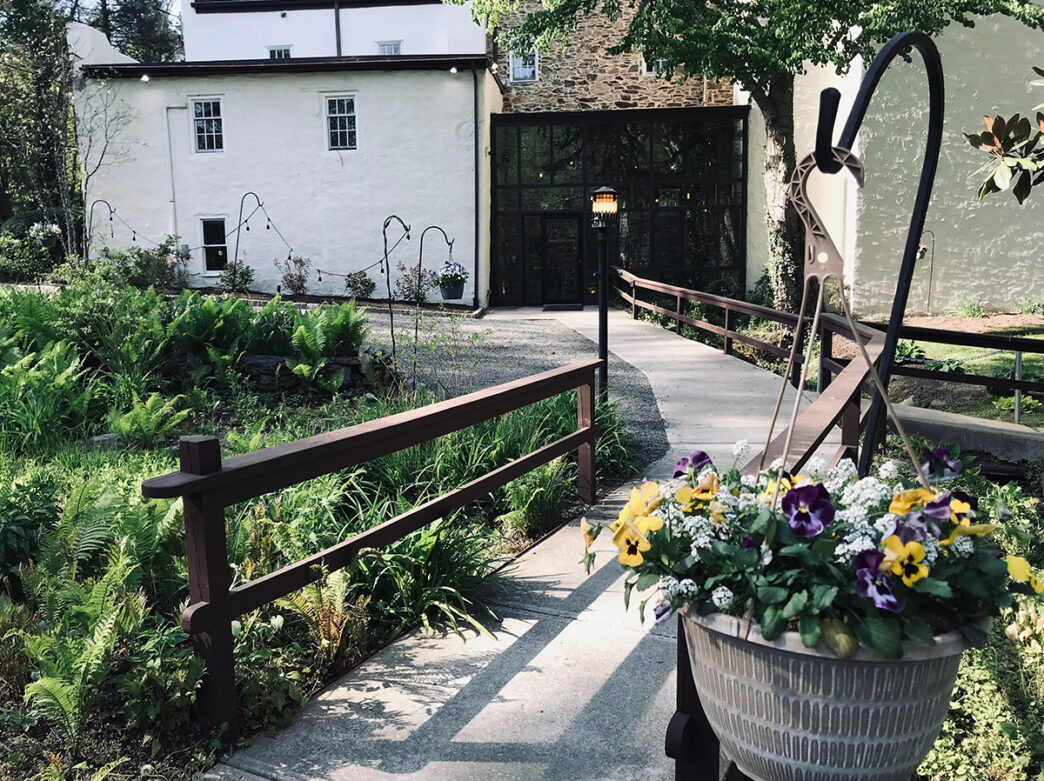A flower-filled garden and walkway lead up to a glass entryway surrounded by historic stone-and-stucco exterior of Hedgerow Theatre Company.