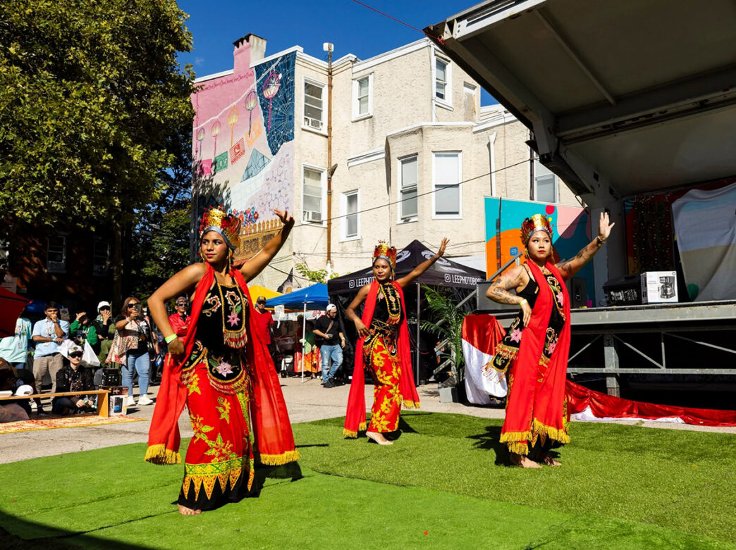 Three dancers in colorful traditional attire perform outdoors during an Indonesian cultural festival, with spectators, vendor tents and a vibrant mural behind them.