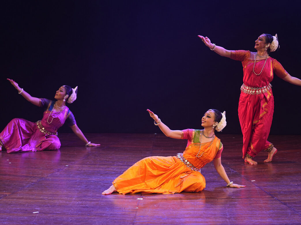 Three dancers in vibrant orange, pink and red traditional costumes performs expressive choreography onstage during a dance performance.