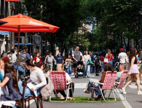 Crowds stroll along a car-free Walnut Street during Open Streets: West Walnut, while visitors dine beneath umbrellas and relax in lawn chairs set up in the roadway.