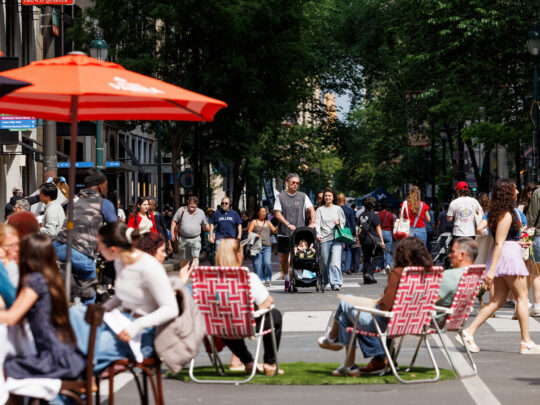 Tijdens het evenement ‘Open Streets: West Walnut’ slenteren mensen over de autovrije Walnut Street, terwijl bezoekers onder parasols dineren en ontspannen in tuinstoelen die op de rijbaan zijn neergezet.
