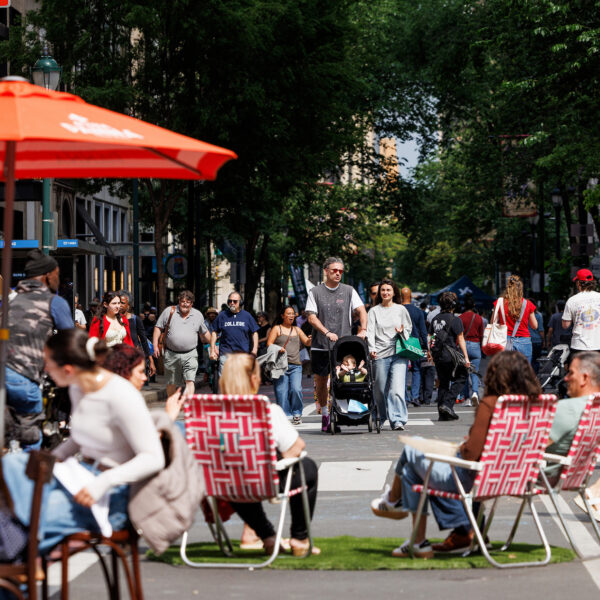 Crowds stroll along a car-free Walnut Street during Open Streets: West Walnut, while visitors dine beneath umbrellas and relax in lawn chairs set up in the roadway.