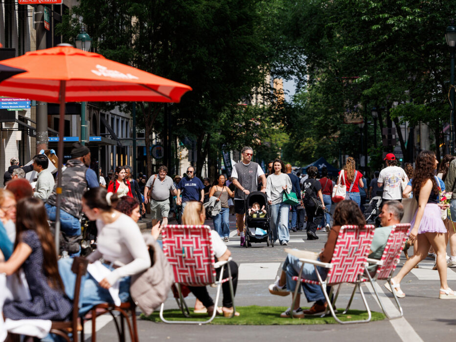 Crowds stroll along a car-free Walnut Street during Open Streets: West Walnut, while visitors dine beneath umbrellas and relax in lawn chairs set up in the roadway.